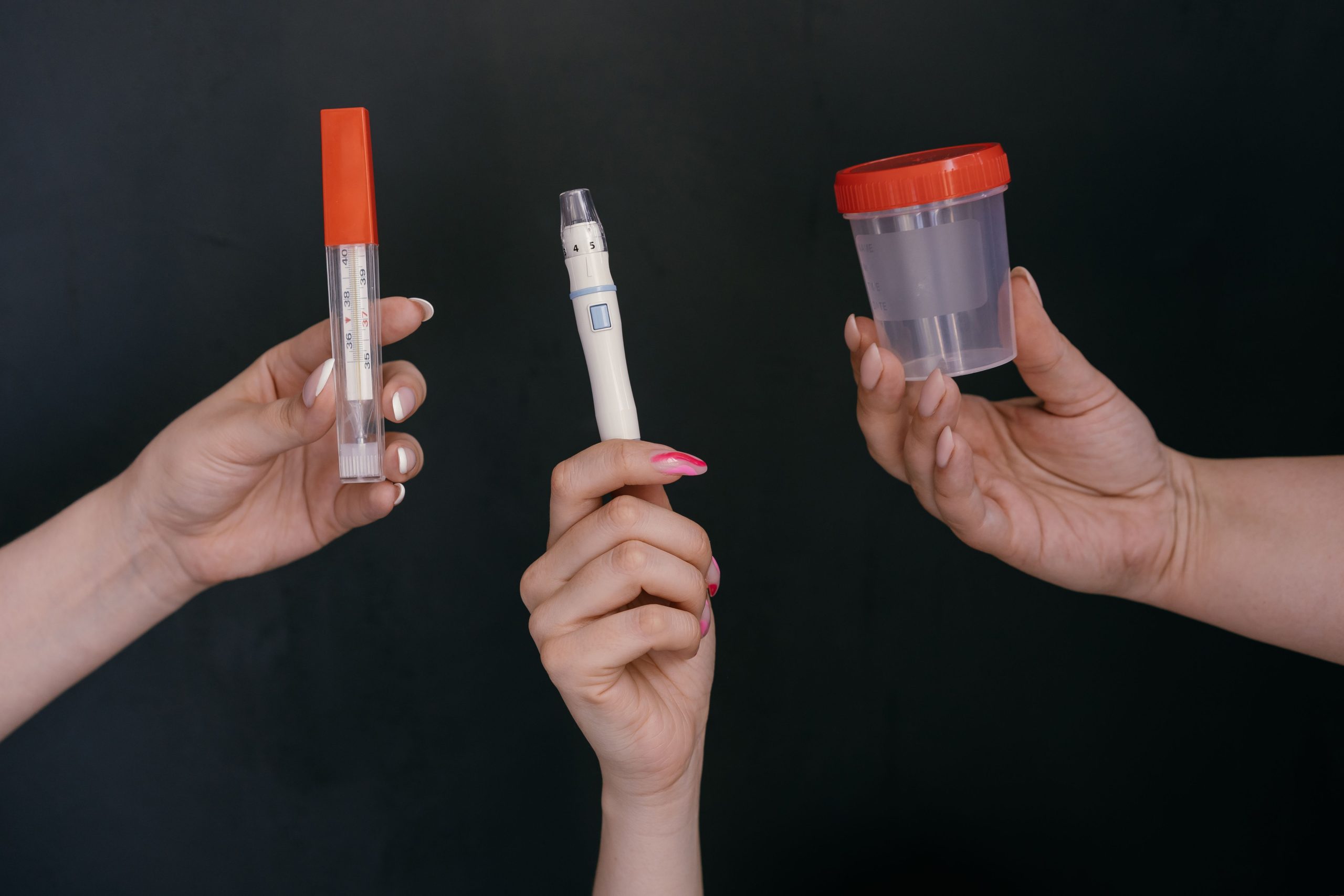 Three hands holding medical tools including a thermometer, lancet, and specimen container.